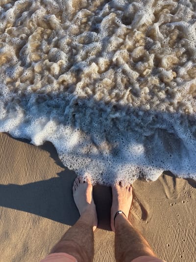 A first-person perspective captures feet standing on a sandy beach where foamy waves are rolling in. The water is a textured mix of tan and white bubbles. Smooth, tan sand surrounds the water's edge and contrasts with the rougher texture of the wave. Visible are the lower legs of a person wearing a light pink shirt. One ankle has a black anklet. Shadows of the person are also cast onto the dry sand.