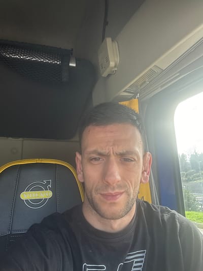 A sole adult male is depicted in a selfie taken within the interior of a Volvo truck cab, located in Tortoreto, Italy. The subject, possessing short dark hair and light stubble, gazes directly at the camera with a neutral to slightly furrowed expression. He is attired in a black t-shirt, displaying a partially visible white graphic logo on its front, consistent with Emporio Armani branding. To the left of the subject, a black seat headrest with yellow trim and a prominent yellow 'VOLVO' emblem is visible. Overhead, a cream-colored, rectangular device, identified as a carbon monoxide or smoke detector, is affixed to the ceiling panel above the subject's right. A black mesh storage net is mounted above the bunk area to his left. Through the truck's passenger-side window, strong daylight illuminates an exterior scene featuring green vegetation, a distant utility pole, and a roadside embankment, indicating the vehicle is stationary.