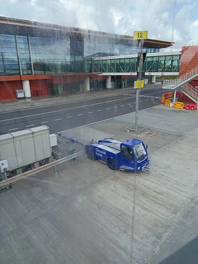 The image depicts an airport scene in Jaipur Municipal Corporation, India. From an elevated viewpoint, possibly from an airplane window or a higher floor of the terminal, the scene includes a blue ground vehicle with the words "Ground Force" visible. The vehicle is parked on the concrete apron adjacent to a building with large glass windows. A yellow sign with the number "12" is visible near the building. Additional equipment or structures are present near the ground vehicle. The environment includes a road or pathway with white lane markings. Luggage carts are parked near the building's elevated pedestrian bridge. The sky is visible with some cloud cover.