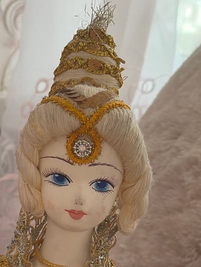 A close-up shot focuses on the face and upper body of a doll, likely an ornament. The doll has a pale complexion, blue eyes with long painted lashes, and rosy cheeks. Its lips are painted a bright red. The doll wears an elaborate headdress and necklace, both adorned with gold and silver thread, sequins, and a circular, jeweled centerpiece on its forehead. The doll's hair is blonde and styled in braids. The background is blurred, showing a soft pinkish-beige textured fabric to the right and a lighter, possibly sheer fabric with a subtle pattern to the left. The overall lighting is soft and diffused. There is no indication of the specific location within Lyambir', Russia, as the image is of an indoor object with a neutral, out-of-focus background.