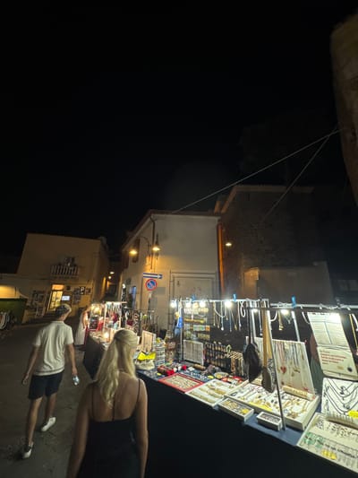 The image depicts a nighttime scene at a street market in Orosei, Italy. In the foreground, a woman with blonde hair, wearing a black dress, walks away from the camera. To her left, a man with short, curly hair, wearing a white polo shirt and black shorts, walks ahead of her. They are on a cobblestone street lined with buildings.
The market stall in the center features a variety of jewelry, including necklaces, bracelets, and earrings displayed on racks and in trays. Lights illuminate the stall, casting a warm glow on the merchandise. In the background, more market stalls and buildings are visible, with warm lighting emanating from some of the structures. The overall atmosphere suggests a lively evening event in a historic Italian town.