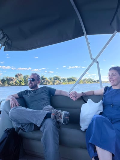 A couple is sitting together on a boat on the Zambezi River in Livingstone, Zambia. The man is wearing a dark green shirt, gray pants, and sunglasses. The woman is wearing a blue dress. They are holding hands. A canopy overhead provides shade. The landscape features trees and a blue sky with clouds.