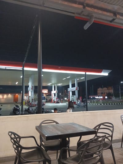 This image depicts a nighttime scene at a gas station, possibly in Meunasahceubrek, Indonesia. The gas station, illuminated by bright lights, features several fuel pumps under a large canopy. A KFC restaurant is visible adjacent to the gas station. In the foreground, a dark-colored table and chairs suggest an outdoor seating area, perhaps for a cafe or restaurant associated with the gas station.

Several motorcycles are parked near the gas station. In the background, traffic and buildings can be seen, indicating an urban or semi-urban environment. A young boy is partially visible in the middle ground, looking towards the gas station. Other individuals and vehicles are also present but are not clearly distinguishable due to the distance and low light conditions.

The time of day is clearly night, as indicated by the dark sky and the artificial lighting. There are no immediate cues about the weather, but the scene appears dry. The overall mood is one of a functional, active commercial area at night, with the lights of the gas station and KFC creating a beacon in the darkness.

Visible text includes "HARVEST" on a building on the left, with the tagline "BEST WASTE. NO HUNGER." The KFC logo is also clearly visible. Another sign displays "MAAF PELAYANAN DIBATASI" which translates to "SORRY, SERVICE IS LIMITED". One of the gas pumps has "PERTAMINA DEX" written on it, which is a brand of fuel in Indonesia.