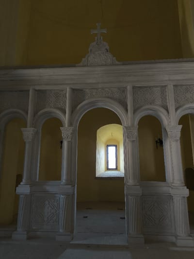 The image displays the interior of a religious structure, likely a church or chapel, situated within the Khotyn Fortress complex in Khotyn, Chernivtsi Oblast, Ukraine. The scene is static and devoid of human presence.

A prominent, multi-arched architectural screen, appearing to be an iconostasis or altar barrier, dominates the foreground and mid-ground. This structure is crafted from light-colored, possibly white or grey, carved stone or stucco. It features a series of rounded arches supported by pilasters or columns with ornate, scrolled capitals. The spandrels and lower panels of the structure are adorned with intricate relief carvings, exhibiting geometric and possibly stylized floral patterns. Centrally, atop the screen, a decorative pediment-like element is crowned by a stylized cross.

The walls beyond this screen are uniformly painted in a pale yellow or ochre hue. A single, tall, narrow, and deeply recessed rectangular window with a dark frame is visible in the background, emitting bright natural light into the space. The floor is composed of large, light-colored flagstones or tiles. A dark, wall-mounted fixture, possibly a sconce or candelabra bracket, is partially visible on the right wall.
