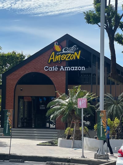 This is a street-level, eye-level, slightly wide-angle photograph of a "Cafe Amazon" establishment located in Ban Ko Pru, Thailand. The cafe building is constructed with red brick, featuring a dark, gabled roof. Prominently displayed on the facade are two signs. The upper sign features the "Cafe Amazon" logo with a parrot illustration and the text "Cafe Amazon" in yellow. Below this, a white sign reads "Café Amazon". An arched entrance leads into the cafe, with steps ascending to it.

Several elements contribute to the environment:

*   **Vegetation:** Palm trees and other leafy trees are visible around the cafe, with one large tree on the right side of the frame partially obscuring the building.
*   **Signage:** In addition to the cafe's signs, a green vertical banner with the "Cafe Amazon" logo is placed to the left of the entrance. Another similar banner is visible on the right, near a lamppost. A pink sign with a female figure and a 'P' indicating a parking space for women is also present.
*   **People:** A person in a blue shirt and black pants is walking past the cafe on the right side of the image.
*   **Objects:** A lamppost stands in front of the cafe. A trash can with a "Cafe Amazon" logo is partially visible on the left. A white car is partially visible in the lower right corner. The ground is paved with concrete tiles.
*   **Atmosphere:** The sky is blue with scattered white clouds, suggesting daytime. The lighting is bright, indicating sunlight.

The scene captures a typical commercial streetscape in Thailand, with the prominent branding of Cafe Amazon.