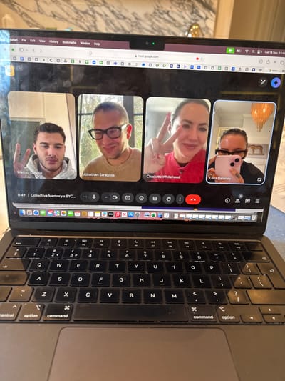A dark-colored laptop, likely a MacBook with a UK QWERTY keyboard, displays an active Google Meet video conference, observed in the Royal Borough of Greenwich, United Kingdom. The laptop's webcam exhibits a green active light. The screen, showing some smudges, presents a Google Chrome browser window, with the URL "meet.google.com" visible. The system date "Tue 18 Nov" is displayed in the top right corner.
The Google Meet conference, titled "Collective Memory x EYC...", features four participants arranged in a grid:
1. **Matteo Gaggia:** A male with short dark hair, wearing a light grey hoodie, making a peace sign with his right hand. His background is a light-colored indoor wall.
2. **Jonathan Saragossi:** A male with short hair and glasses, wearing a light-colored sweater, smiling. His background shows outdoor foliage.
3. **Charlotte Whitehead:** A female with dark hair pulled back, wearing a red top, making a peace sign with both hands. Her background is indistinct.
4. **Cora Delaney:** A female with dark hair pulled back, wearing glasses and a dark top, holding a pink smartphone, with its camera lens visible. Her background includes an ornate light fixture and a framed image of a horse.
The conference time 11:49 is displayed on the bottom left of the meeting interface, which also shows controls for microphone, camera, captions, hand raise, and ending the call. The laptop is situated on a light-colored surface. A blurred indoor background, including a decorative item and a light-colored wall, is visible behind the laptop screen.