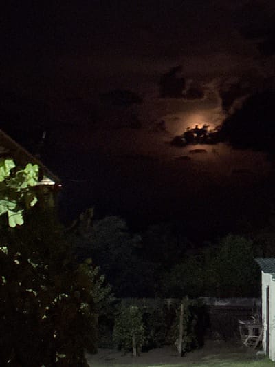 The image depicts a night scene in Serhiivka, Ukraine, with a full moon partially obscured by clouds. A building with a white exterior and a dark roof is visible on the right side of the frame. In the foreground, there is dense foliage, likely trees and bushes, with some illuminated leaves. A wooden fence runs through the middle ground, separating what appears to be a yard or garden area from the background. A piece of outdoor furniture, possibly a chair or small table, is situated near the fence on the right. The overall ambiance is dark and quiet, with the moonlight casting a faint glow on the scene.