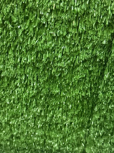 The image shows a close-up of artificial turf, densely packed with short, vibrant green blades. The texture is slightly irregular with subtle variations in shading, giving it a somewhat realistic appearance. Light reflects off the plastic-like fibers, adding a sheen to the surface. Overall, the focus is on the texture and color of the synthetic grass, providing a tactile and visual representation.