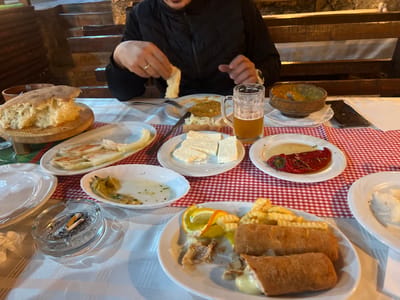 The image depicts a dining scene inside a rustic restaurant or tavern in Despotovac Municipality, Serbia. One male individual is seated at a table, visible from the chest down. He wears a dark, possibly quilted, jacket and a ring on his left ring finger. His hands are actively engaged with food, specifically tearing a piece of bread.
The table is covered by a white tablecloth, with a red and white checkered runner laid over it. Numerous dishes are present: a large, partially torn loaf of rustic bread on a wooden board; a plate with rectangular slices of white cheese; plates containing baked or fried peppers (one red, one yellowish-green); a plate with a spread; a plate with light-colored, flat food items; and a main course plate featuring two large, breaded and fried rolls (one partially cut revealing a creamy filling), a portion of crinkle-cut french fries, a lemon wedge, and an orange slice. A small, ornate ceramic bowl contains a yellowish dish, possibly a bean stew.
A clear glass mug filled with an amber liquid, consistent with beer, sits on the table. An ashtray containing two cigarette butts and ashes is visible. A dark-colored smartphone lies face down on the tablecloth. Additional empty white plates, silverware, and a crumpled paper napkin are also on the table.
The background shows warm, dim lighting illuminating wooden benches and structural elements, including a set of wooden stairs on the left, indicative of an interior dining establishment.