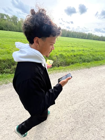 A person with curly dark hair, wearing a black jacket over a white hooded sweatshirt, black pants, and green and white shoes, is standing on a gravel path. The person is holding a black smartphone in their right hand and looking at the screen. A carton of juice sits on the grassy area to the left. The background consists of a green field with trees along the horizon and a cloudy sky. The photo was taken near Chelles, France.