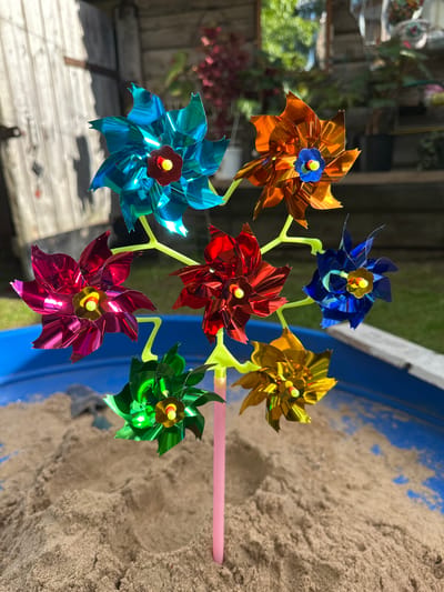 The image displays a colorful pinwheel with seven individual pinwheel flowers. The pinwheels are attached to a lime green stem, which is inserted into a pink tube, presumably a handle. The pinwheels themselves are made of metallic material and come in various vibrant colors: orange, blue, red, gold, light blue, green, and a deep reddish-pink. Each pinwheel has a small yellow and gold center.
The pinwheel is positioned outdoors, with a background that suggests a backyard or garden setting. To the left, there's a wooden fence and some greenery, possibly plants or trees, with some out-of-focus elements that might be garden decorations. The right side of the image shows a blue container filled with sand, suggesting it might be a sandbox or a part of an outdoor play area. The sunlight indicates it is daytime, and the shadows cast by the pinwheel suggest a bright, sunny day.
There is no visible text in the image. The overall mood is cheerful and playful, evoking a sense of summer or outdoor activity. The composition focuses on the pinwheel, showcasing its bright colors and intricate design against the sandy backdrop. The location context of Piriutino, Russia, is provided but not visually evident from the image itself.