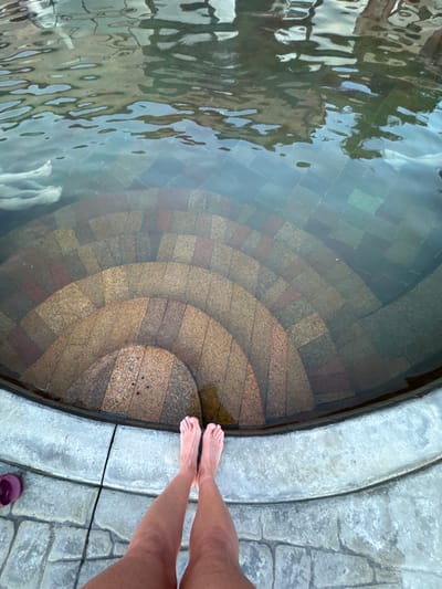 A top-down view of a shallow circular pool with concentric steps descending into the water. The pool is lined with tiled surfaces that appear to change color with depth, from dark greens and blues near the top to warmer browns and reds lower down. The water surface is rippled, reflecting the environment above. Two bare feet and lower legs are visible on the right edge of the frame, suggesting a person standing at the pool's edge. The pool's edge is made of light grey stone, and the surrounding paving is also grey and made up of interlocking blocks. A purple sandal is visible on the bottom right edge of the frame. The photograph was taken in Kara-Oy, Kyrgyzstan.