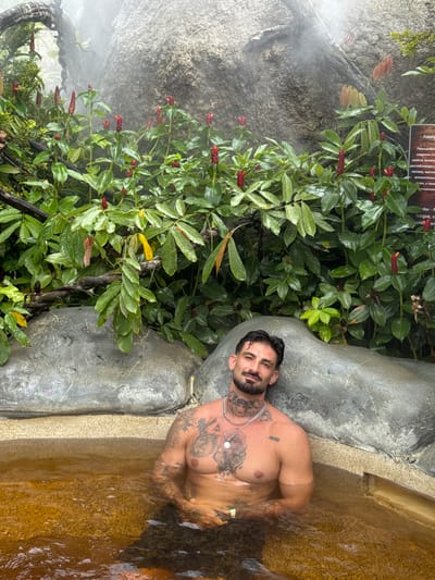 Here is a detailed description of the image:
**Location:** This image appears to be taken at a spa or a natural hot spring, possibly in a tropical or subtropical location. Based on the information provided, it is potentially located in the city of Ban Khlong Khian บ้านคลองเคียน, Thailand.
**People:** The primary subject is a shirtless man with tattoos. He has a beard and appears to be relaxed.
**Environment:** The scene is lush with greenery. The man is in water, possibly a hot spring or bath of some kind. There are large grey stones or concrete structures around the water. Additionally, mist or steam is present, adding to the ambiance of a natural, relaxing environment.
**Subjects and Objects:**
* The man himself with tattoos.
* Water which may be from a hot spring.
* Lush, tropical greenery and red-flowering plants.
* Grey stones or concrete forming the edge of the bath.
* A sign with text on it, possibly containing information about the plants or spa.
* A chain around the man's neck.
**Actions/Interactions:** The man is relaxing in the water, leaning against the side.
Overall, the image depicts a scene of relaxation in a natural spa-like setting, with the man as the central figure enjoying the tranquil environment.
