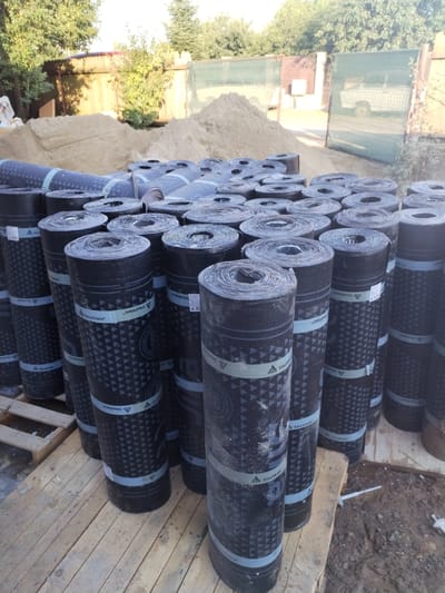 A construction site in Dumbrăveni, Romania, is depicted with several stacks of black roofing rolls, likely bitumen membranes, placed on wooden pallets. The rolls are cylindrical and tightly wound, many of them featuring a light blue band with text and triangular patterns. In the background, piles of sand and construction debris are visible, along with some trees and a fence. The overall scene suggests an outdoor storage area for building materials.