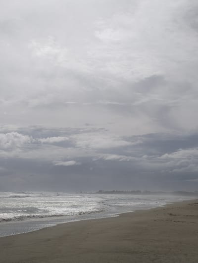 The image depicts a deserted beach under an overcast sky. The foreground shows a sandy shore with subtle imprints, possibly from footprints. The middle ground is occupied by a turbulent ocean with white-capped waves rolling towards the shore. The horizon is blurred by mist or fog, with a faint line of trees visible in the distance. The overall impression is one of a somber and moody seaside atmosphere. There are no people or animals visible in the scene, and no structures or man-made objects are apparent, suggesting a natural and remote location. The location of Meudheun, Indonesia, is not visually identifiable within the image itself, but is provided as context.