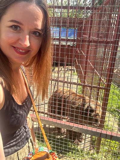 A woman is taking a selfie with a bear in a cage at a zoo. The woman is on the left side of the image, smiling at the camera. She has long, brown hair and is wearing a black tank top and khaki pants. She is also carrying a colorful bag over her shoulder. The bear is on the right side of the image, behind a metal cage. It is a large, brown bear. The background of the image is a zoo enclosure with grass and trees. The location is identified as Medvedevo, Russia.