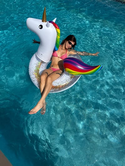 A woman is floating on an inflatable unicorn in a swimming pool in Sant Vicenç de Montalt, Spain. The woman is wearing a red and white checkered bikini and sunglasses. She has tattoos on her arms and legs. The unicorn is white with a rainbow mane and tail. The swimming pool is blue and there are tiles on the bottom. The sun is shining and the water is clear.