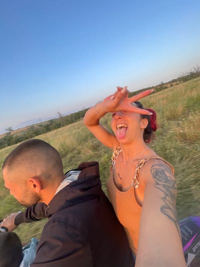 In a field near Zheglartsi, Bulgaria, a man with a shaved head is riding an ATV, with a woman behind him. She is sticking her tongue out, wearing a tan tank top and has tattoos on her arm. The sun is setting, casting a warm glow on the scene.