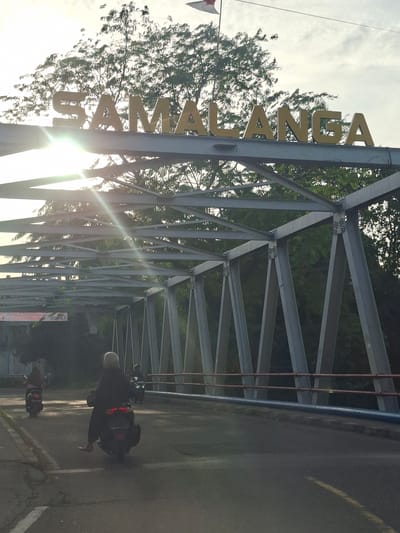 The image shows a bridge over a road in the city of Meurah, Indonesia. The bridge has a large metal sign with the word "SAMALANGA" written in gold letters. The sun is shining brightly, casting rays through the bridge's structure. Several motorcycles are on the road, with riders wearing helmets. In the background, there are trees and a cloudy sky. There is also a flag visible on a pole in the upper right corner. The overall scene appears to be a typical day in the city.