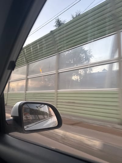 The image shows a view from the passenger side of a car moving along a road, likely in Mstikhino, Russia. A sound barrier, constructed of green corrugated metal panels and translucent inserts, runs parallel to the road. The inserts reflect the sky and trees, and a single yellow light is visible through one of them. The car's side mirror is also visible, reflecting a person holding a phone and the road ahead, which appears wet. The sky is overcast with hints of sunset.