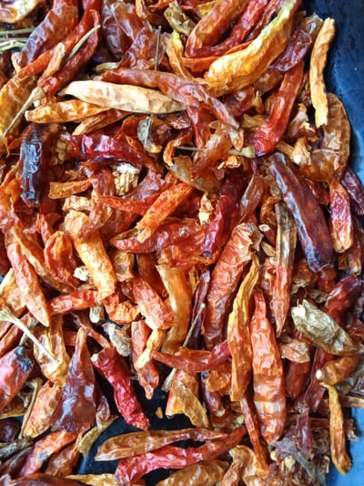 A pile of dried red chili peppers is shown in close-up, filling the entire frame. The peppers vary in color from deep red to a lighter orange and brown, with some appearing shriveled and others more plump. They are arranged haphazardly, overlapping each other. The background is a dark, slightly textured surface. There are no people or any discernible city elements visible in the image. The scene is an indoor or still life arrangement of dried chilies, not a street scene in Dongo, Nigeria.