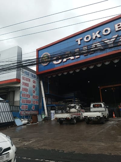 This image depicts a commercial street scene in Cipendawa, Indonesia, under an overcast sky. The primary subject is a large, two-story building on the right with a blue facade, displaying "TOKO BANGUNAN CAHAYA BANGUNAN" (Construction Material Store Cahaya Bangunan) in prominent white lettering. A white banner with scalloped edges hangs below the main sign. The building's large, open front reveals a dark interior filled with stacked materials, suggesting a warehouse or large store.
Adjacent to this building on the left is a smaller structure featuring a blue and white sign that reads "PUSAT PENJUALAN" (Sales Center), listing construction materials such as "BESI BETON," "SPANDEX," "BONDEX," "WIRE MESH," "HOLLOW," and "BAJA RINGAN," concluding with "TERLENGKAP TERMURAH" (Most Complete, Cheapest). Stacks of metal sheets and other construction materials are visible outside this building.
Three white pickup trucks are present. One is partially visible in the bottom left foreground, showing its front hood and bumper (license plate 880 HO). Two additional white pickup trucks are parked in front of the large blue building, facing away from the viewer. The left truck features a metal frame on its bed and a "Solarfruit" sticker (license plate F 8596 RJ). The right truck has an open bed (license plate B 8390 ND). A light-colored scooter or motorcycle is partially visible near the smaller building's entrance.
Numerous black electrical wires are strung horizontally across the upper-mid section of the image. The asphalt ground is wet and reflective, indicating recent rain or damp conditions. On the far right, a single individual wearing a high-visibility vest is partially visible near an orange traffic cone.