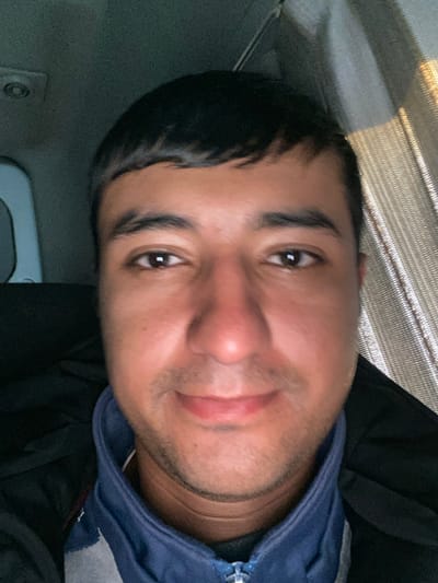 A male individual is depicted in a close-up, front-facing selfie taken inside what appears to be a vehicle in Rabatak, Uzbekistan. The subject has dark, short hair, dark eyes, and a light stubble on his face, which displays a neutral to slight smiling expression with a slightly flushed nose and cheeks. He is wearing a dark blue zippered jacket or top over a grey garment, underneath a dark outer jacket. The background on the left shows a grey vehicle interior, including a circular light fixture on the ceiling. On the right, a textured, light-colored reflective surface, possibly a sunshade or curtain, is visible, illuminated by an external light source. The individual is looking directly at the camera, implying he is actively taking the photograph.