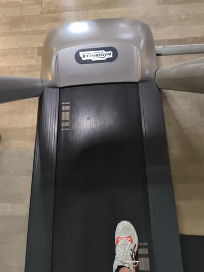 The image shows a person's white and orange athletic shoe on the dark running belt of a Technogym treadmill. The treadmill is positioned on a light-colored wood-look floor. The top console of the treadmill, which features the Technogym logo, is visible, as are the side rails of the machine. The scene appears to be indoors, likely in a gym or fitness facility. The lighting suggests it could be daytime, but there are no direct indicators of the time of day or weather outside. The focus is on the treadmill and the person's foot, implying an activity such as running or walking is taking place or about to take place. The overall impression is one of a fitness-focused environment.