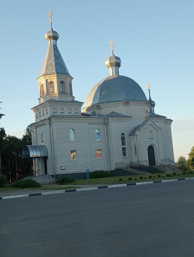 The image shows an Orthodox church, likely located in the Вяйнянскі сельскі Савет, Belarus, given the context. The church is constructed of light-colored bricks and features a prominent central dome and a tall, slender bell tower, both topped with golden crosses. The bell tower has a pointed roof and multiple arched windows. The main body of the church also has arched windows, some with colored glass that reflects light. A set of stairs with black railings leads up to the main entrance, which is a dark-colored door under an arched facade. To the left of the main entrance, a glass-covered porch with a curved roof shelters another entrance. The church is surrounded by green lawn and some small, manicured bushes. In the background, a line of dark green trees can be seen. A paved road with a curb and visible road markings runs in front of the church. The sky is clear and blue, suggesting it is daytime, likely late afternoon due to the warm lighting. There are no people visible in the image.