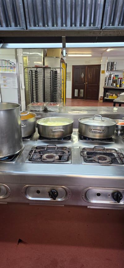 The image shows a commercial kitchen at a restaurant in Sallent de Gállego, Spain. The focus is on a stainless steel stove with four burners, two of which are in use. On the left burner, a large silver pot containing a yellow liquid is cooking. Next to it, on the second burner from the left, is a silver pot with a lid, containing a light yellow liquid that appears to be boiling. To the right of these, two more burners are visible, one with a silver pot with a lid, and the last one appears to be off. The stove has control knobs below the burners. In the background, a stainless steel rack holding trays covered in plastic wrap, containing raw meat, is visible. Further in the background, there are double wooden doors and kitchen equipment. The overall environment is that of a busy, functional kitchen.