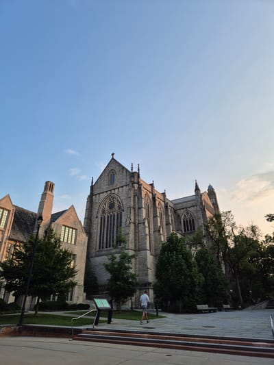 This is a photograph of Graduate College at Princeton University in Princeton, New Jersey, USA. The image depicts the gothic architecture of the college buildings, characterized by stone construction and large, arched windows. The main building features a prominent, tall central structure with a steepled roof and spire, adorned with intricate stonework and decorative elements. To the left, another section of the building, with a more rectangular facade, has a tall, square chimney and dormer windows.

The buildings are surrounded by mature trees with lush green foliage, casting shadows on the surrounding paved walkways and grassy areas. In the foreground, a person is walking away from the camera, wearing a light-colored shirt and shorts. The sky is a clear blue with a few wispy clouds, and the lighting suggests it is either early morning or late afternoon, with the sun casting a warm glow on the stone structures. There are benches visible in the background, indicating a public space.