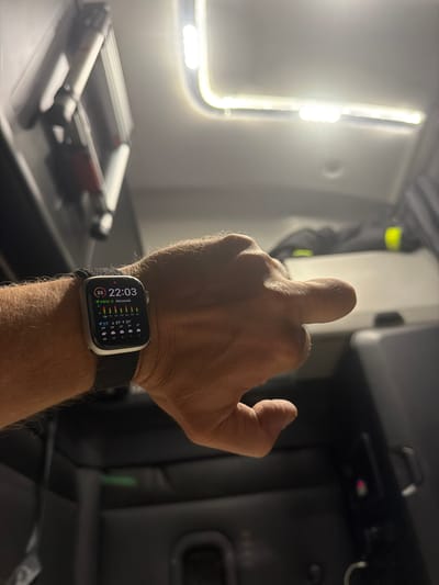 An individual's left wrist, adorned with a dark fabric watch band and a smartwatch, is positioned in the foreground. The smartwatch displays the time as 22:03 and features various weather-related icons and data. The background suggests the interior of a vehicle, possibly a truck cab, with a bright light source overhead and dark upholstery visible. The scene appears to be dimly lit, indicating a nighttime or low-light environment. There are no visible landmarks or indicators to place this image in New Britain, United States of America.