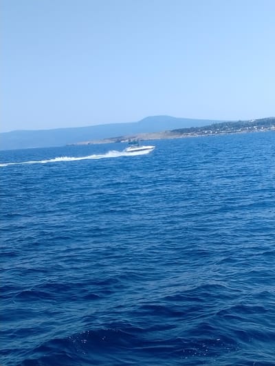 A speed boat with a white hull is captured in motion on the deep blue Adriatic Sea. The boat is leaving a prominent white wake behind it as it travels from left to right across the frame. In the background, a hazy blue mountain range forms the horizon, with a strip of coastal land dotted with buildings visible in front of it. The sky is a clear, pale blue, indicating a bright, sunny day. The foreground is dominated by the textured surface of the sea, with small waves creating a pattern of light and shadow. The scene depicts a typical summer day on the coast of Šilo, Croatia.