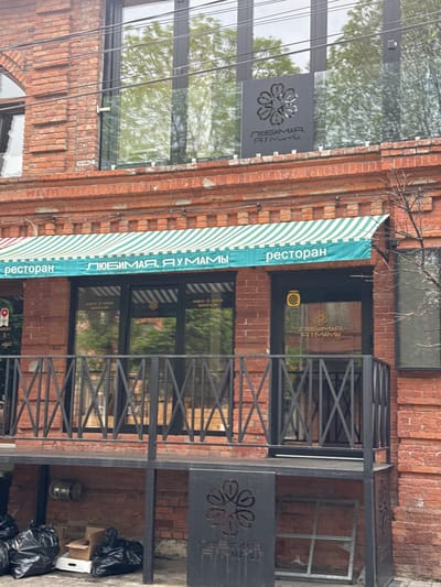 The image depicts the exterior of a restaurant called "Lyubimaya, Dumami" ("Beloved, Dumami") located in Akhieli, Georgia. The restaurant is housed in a brick building with a striped awning. The signage includes the name of the restaurant in Russian, accompanied by a floral logo. A black metal structure, possibly a decorative railing or small porch, is present in front of the entrance. Trash bags and cardboard boxes are visible near the base of the structure. Power lines run along the top of the building.