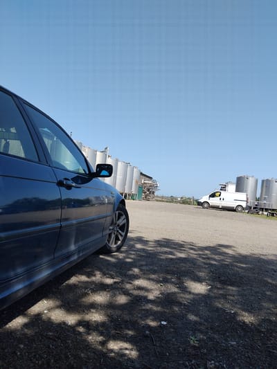 The image shows a dark blue car parked on a gravel surface adjacent to a complex of industrial tanks and piping, with a white van parked further along the same side. The sky is clear and blue. Tree shadows are cast on the ground next to the car. The location is likely in the city of Stafford, United Kingdom, based on the prompt.
