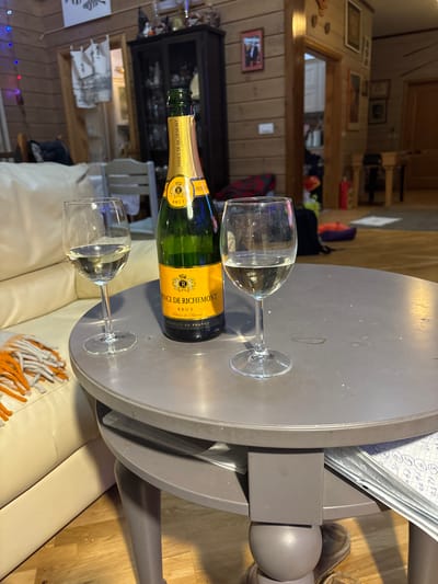 A bottle of Prince de Richmont Brut lies horizontally on a gray, round table. Two empty wine glasses flank the bottle. The table sits atop a white couch with a fringed throw. In the background, the interior of a home can be seen, including wooden walls, a hutch, and various decorations. A notebook is on the table next to the bottle.