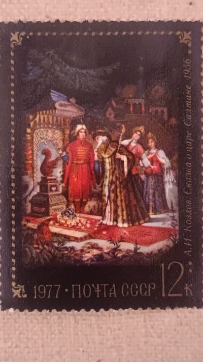This is a postage stamp depicting a scene from a fairy tale. In the foreground, four figures in ornate, historical costumes are gathered around a red carpeted area. The central figure, an older person with a long, striped robe and a staff, appears to be the focal point. To their left, a person in a vibrant red coat and matching hat stands prominently. To the right of the central figure, two other individuals, one in a red and white ensemble and another in a dark gown with red accents, are also present.

Behind the figures, a rich, patterned screen or tapestry forms the backdrop, suggesting an indoor, opulent setting. To the far left, a pedestal holds a red, bird-like statue, possibly a squirrel, and a dark, ornate structure. The overall scene is richly colored with deep reds, browns, and blues.

The stamp has a dark border with decorative elements along the edges. Visible text on the stamp includes "1977", "ПОЧТА СССР" (which translates to "Post of USSR"), the number "12", and the letter "К". Along the right edge, there is text in Russian: "А.И. Козлов. Сказка о царе Салтане 1956", which translates to "A.I. Kozlov. The Tale of Tsar Saltan 1956". This indicates the stamp commemorates the 1956 tale by A.I. Kozlov, likely illustrating a scene from it, and was issued in 1977. The imagery suggests a formal or ceremonial gathering within a historical or fictional court. No specific time of day or weather is discernible. The location context of Onaldy, Turkmenistan is not visually represented on the stamp itself.