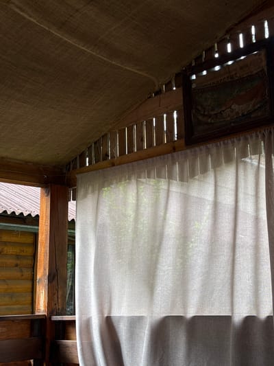 The image presents the interior of a rustic, wooden semi-open structure, identified as being in Zviahel, Ukraine. No individuals are depicted. A light-colored, semi-translucent fabric curtain, possibly linen, occupies the majority of the right and lower-middle sections, exhibiting vertical folds and allowing diffuse natural light to filter through, revealing blurred green foliage beyond. Above the curtain, wooden vertical slats forming part of the structure permit narrow shafts of bright sunlight to enter. A dark, woven fabric, consistent with burlap or similar material, is draped across the upper ceiling, forming a canopy. To the left, a robust, rough-hewn wooden post stands vertically, adjacent to horizontal wooden paneling. Beyond this opening, a corrugated metal roof is visible under a bright sky. A framed picture is suspended from the wooden wall above the curtain; its details are largely obscured by shadow and reflection, though inverted Cyrillic text is partially visible on the upper section, and a muted, multi-colored image, possibly a landscape or abstract design, is present below. A thin wire is horizontally strung across the top of the curtain's suspension point. The environment suggests an outdoor or semi-outdoor setting, characterized by natural wood construction and diffused natural lighting.