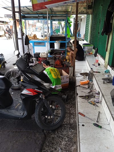 The image shows a scene at a motorcycle repair shop located in Kauman, Indonesia. A black Honda Beat scooter is prominently displayed in the foreground. A man wearing a green shirt is working on a mechanical component, positioned near a red REPSOL box and an air compressor. Another person is seated on an elevated platform along a wall, using their phone. Tools and bottles are scattered on the platform. In the background, a food cart or stall is visible. A second motorcyclist is seen on the road in the distance. The shop is cluttered with various equipment, spare parts, and supplies.