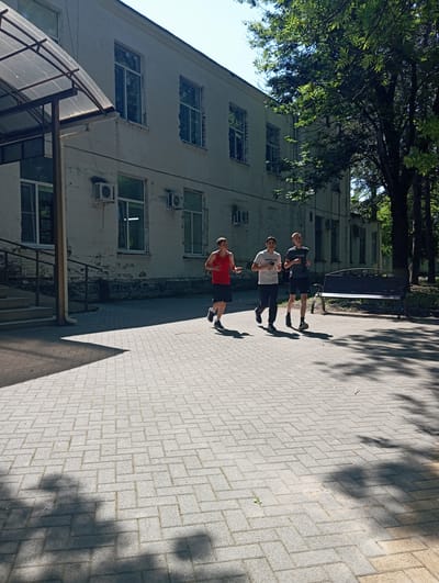 Three young males are jogging on a paved walkway in front of a white, two-story building. The building has several windows and air conditioning units visible. A covered walkway with a transparent roof extends from the left side of the building, and stairs lead up to it. Trees with lush green foliage are on the right side of the frame, casting shadows on the walkway. A dark metal bench is positioned near the trees. The walkway is constructed of rectangular pavers laid in a herringbone pattern. The overall environment suggests an outdoor setting in a park or school campus, possibly in Otradnaya, Russia, given the architectural style and vegetation. The time of day appears to be daytime with bright sunlight creating strong shadows.