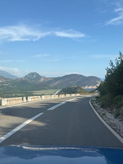 The image captures a winding asphalt road in Grabovica, Montenegro, on a clear day with a blue sky and scattered white clouds. The road features double white dividing lines and a white edge line. A low, weathered stone wall borders the road on the left, with some vegetation and small white structures visible beyond it in a valley. Rolling green hills and rugged mountains form the backdrop, with a glimpse of the sea and a cliffside visible on the right. Dense green trees and foliage line the right side of the road. The foreground shows the reflection of the landscape on the blue hood of a vehicle. There are no people or animals visible in the frame.