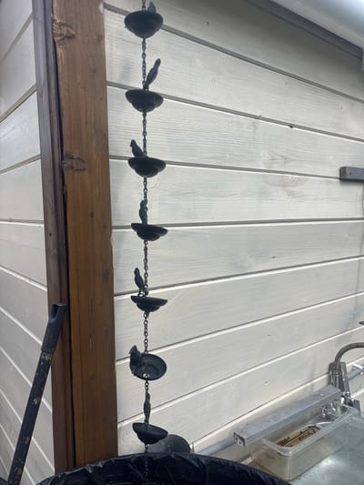 The image shows a close-up of a chain with decorative bird-shaped elements hanging against a white wooden wall. The chain is made of dark metal, with multiple cups and small bird figurines attached to it. To the left of the chain is a thick, weathered wooden post. The background is a horizontal planked wall, painted white. In the lower right corner, a sink or basin with some objects inside is visible, along with plumbing fixtures. The overall scene appears to be outdoors, possibly in a yard or on a patio, given the wooden structures and outdoor elements. The specific location of Лотос Снт (марьино), Russia, is provided as context but not visually depicted in the image itself.