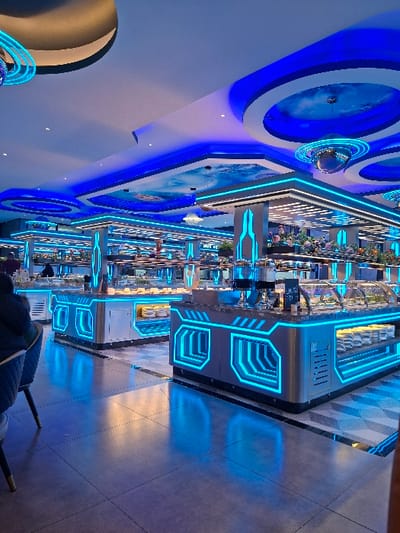 A futuristic-themed restaurant features a long buffet line with food under glass displays. Blue LED lighting outlines the buffet structure and ceiling designs. Decorative, illuminated planet-like objects adorn the ceiling. The flooring has a geometric pattern reflecting the blue light. Modern chairs are visible at tables near the buffet.