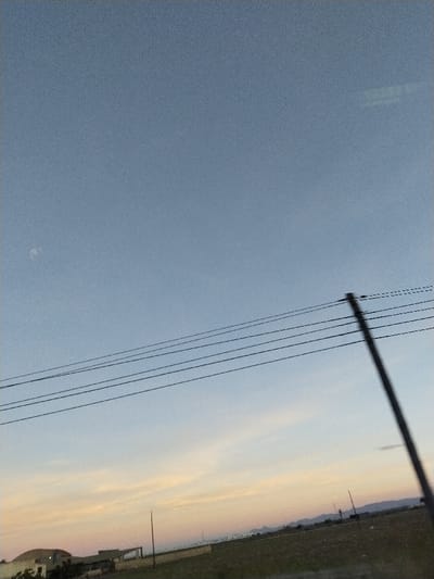 The sky is light blue with hints of orange near the horizon. Power lines stretch across the middle. A blurred pole stands upright on the right. In the distance, low buildings and possibly mountains are visible. The foreground shows a grassy field. The overall image has a slightly grainy or hazy quality.