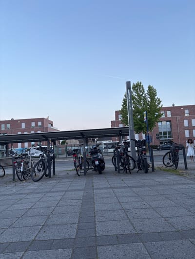 This is a photograph taken at the train station in Gronau, Germany. In the foreground, a paved area with square gray tiles leads to a covered bicycle rack filled with parked bicycles and a scooter. A woman in a white dress walks away from the camera on the right side of the image. Behind the bike rack, a bus is visible, and beyond that, a modern brick building with multiple windows stands under a clear blue sky with a faint contrail. The overall scene depicts an urban environment at what appears to be late afternoon or early evening, given the soft lighting.