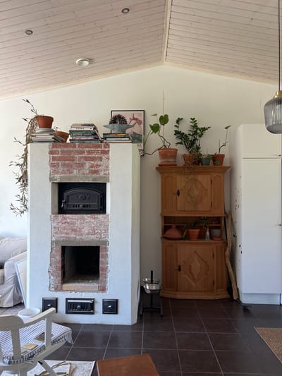 The image shows the interior of a room, likely a living area, featuring a white tiled oven with exposed red brick detailing. The oven has a dark metal door with ornate detailing. Above the oven, a stack of books and several potted plants are visible. To the right of the oven, a tall, light-colored wooden cabinet with decorative carvings holds more potted plants on its top. A white refrigerator is positioned to the far right of the cabinet. In the foreground, a light-colored chair with blue and white striped cushions is partially visible. The floor is tiled in dark brown squares. The room has a light-colored, sloped ceiling made of wooden planks, with visible light fixtures. The scene appears to be a domestic interior in Hämeenlinna, Finland. No people are present in the image, and no actions or interactions are taking place.