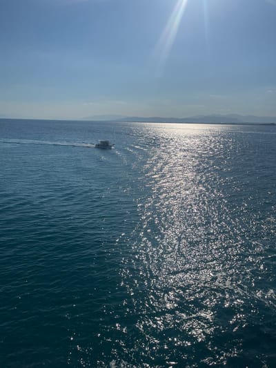 The image depicts a serene seascape under a clear, bright sky, likely during daytime. The sun's glare creates a shimmering effect on the rippling blue water, suggesting a calm and pleasant weather condition. In the distance, a faint outline of mountains or hills can be seen along the horizon, hinting at a coastal location.

A small boat is the only discernible object in the water, leaving a subtle wake as it moves. The boat appears to be a vessel of some sort, possibly a fishing boat or a small ferry, with a light-colored structure. The overall atmosphere is one of tranquility and vastness, with the expansive sky and sea dominating the frame.

The image is oriented vertically, with the sun's rays emanating from the left side, casting a bright glow and creating a sense of depth. The water's surface is textured with numerous small waves, reflecting the sunlight in a dazzling display. The distant landmass is softly blurred, contributing to the sense of distance and peacefulness. There is no visible text in the image. The scene evokes a feeling of calm and openness, characteristic of a peaceful day at sea. Given the location context of Kastro - Kyllini, Greece, the scene is likely set in the Ionian Sea.