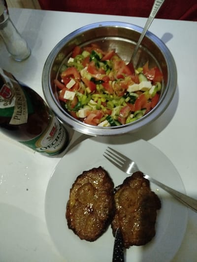 The image depicts a meal on a white table, possibly in Kitzingen, Germany. In the upper portion of the image, a stainless steel bowl contains a chopped salad composed of tomatoes, cucumbers, white cheese cubes, and sliced green onions. A fork is partially submerged in the salad. To the left of the bowl, a brown glass bottle of Pilsener beer, with a green and white label, is visible. In the lower portion of the image, a white plate holds two pieces of cooked meat. A fork rests on the plate, pointing towards the right, and a knife with a black and white patterned handle is placed in front of the meat. The lighting suggests an indoor setting, possibly a home or restaurant.