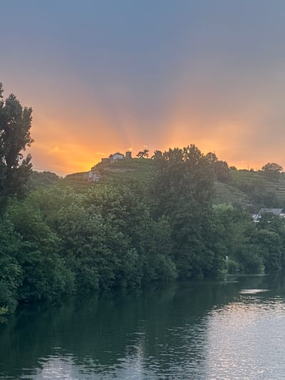 The image shows a serene sunset over the Neckar River in Ludwigsburg, Germany. The sky is a gradient of soft blues and oranges, with the sun's rays peeking through the clouds behind a distant hilltop. On the hilltop, there is a small structure, possibly a castle or an old ruin, surrounded by vineyards. Lush green trees line the riverbank in the foreground, their reflections shimmering on the calm water. The overall scene evokes a peaceful and picturesque atmosphere, typical of a European countryside. The perspective is from across the river, looking towards the hill and the sunset. There are no people or animals visible in the frame.