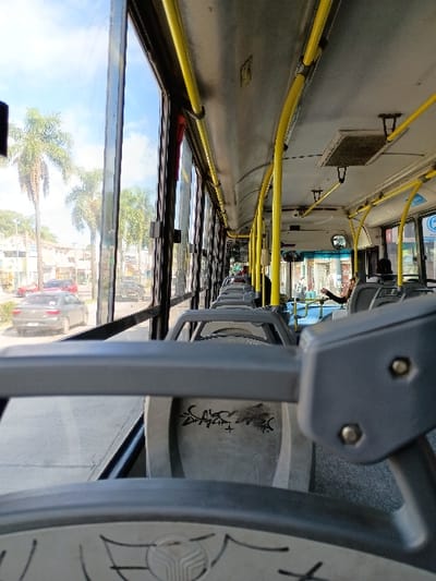 The image shows the interior of a public bus in General Pacheco, Argentina, at a location with coordinates -34.46 latitude, -58.634 longitude. The bus is facing forward with the windows to the right and front of the bus visible. The view from the windows shows the street and surrounding area, including buildings and vegetation. Several empty seats are visible. The seat in the foreground is gray plastic with graffiti on it. Several empty seats are visible in the background, some of which are in close proximity to the front of the bus. A small figure can be seen in the distance sitting in one of the front seats, and another person is leaning over a seat in the back. Several yellow support poles can be seen running from the ceiling to the floor on the right side of the bus. A grey overhead handle is in the foreground and is partially obscured by the seat in front of it. The image is likely taken from the perspective of a passenger sitting in the middle of the bus facing the front. The bus is in motion.